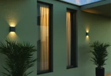 Fachada de uma casa com parede pintada de verde, duas arandelas pretas e dois vasos de plantas.