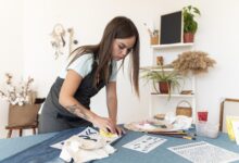 Mulher realizando a pintura de uma calça jeans, sobre uma mesa com materiais de pintura.