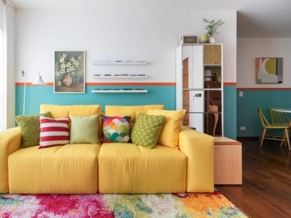 Pintura para sala branca com meia parede azul com faixa laranja, sofá amarelo com almofadas e tapete coloridos.