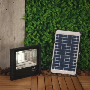 Imagem de um refletor solar preto ao lado de uma placa solar pequena. Ao fundo temos uma parede de plantas artificiais.