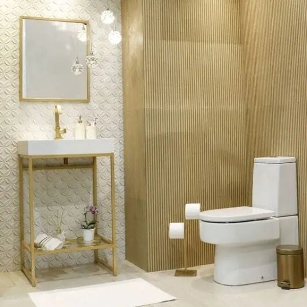 Banheiro com parte da parede com textura, e o restante em dourado. Piso branco e acessórios dourados.