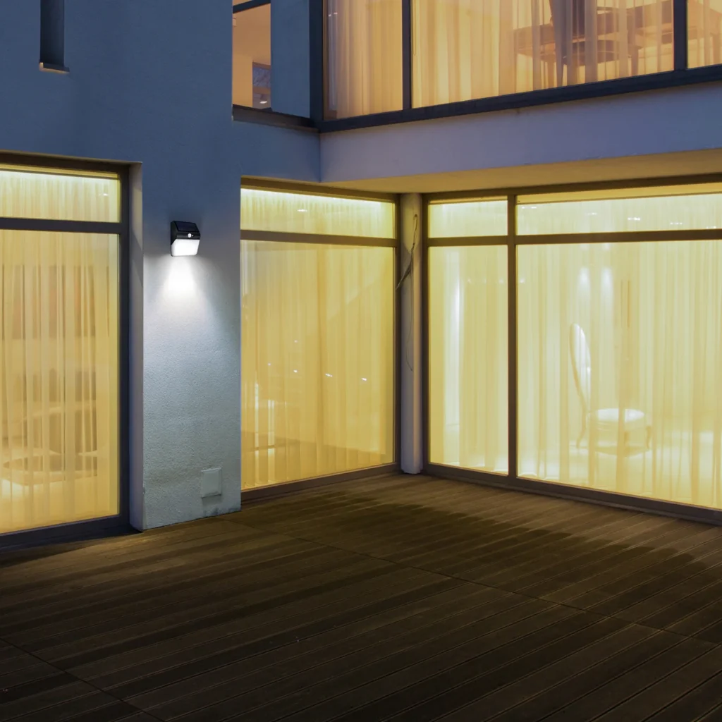 Parede externa com vidros iluminada por uma arandela solar preta de luz branca.