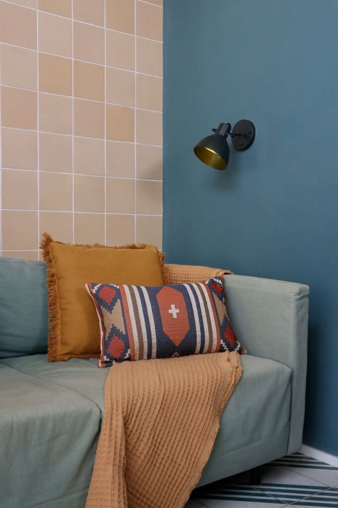 Sofá cinza encontado em uma parede azul, com duas almofadas, sendo uma laranja e outra estampada, em cima de uma manta laranja.