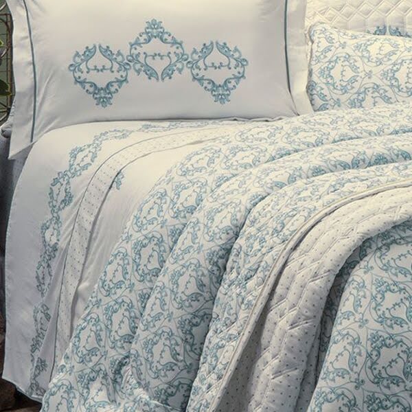 Imagem de uma cama de casal coberta por um edredom estampado com arabescos verde-azulados e branco.