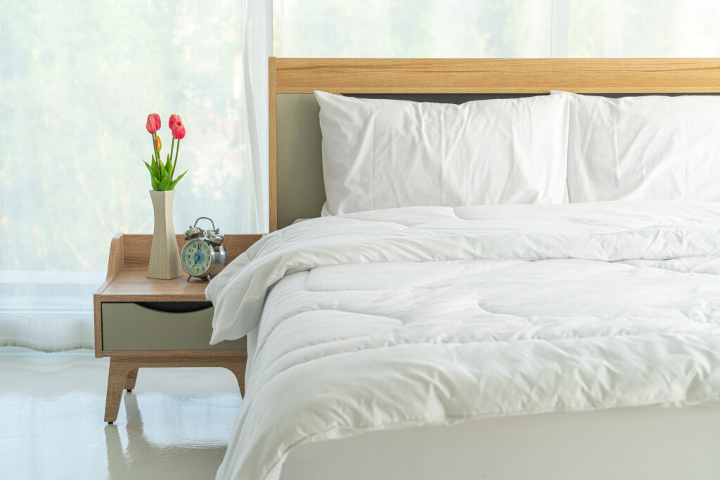 Imagem de um quarto contendo uma cama de casal coberta por um edredom branco, além de uma mesa de cabeceira, um vaso de plantas, travesseiros e um relógio.