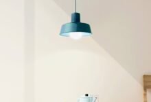 Luminária azul com lâmpada acesa sobre uma mesa pequena de madeira com fruteira, bule e um copo de vidro sobre ela.