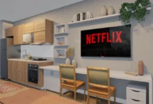 Projeto de studio com parede clara, mesa encostada na parede com duas paredes, uma televisão acima dela e nichos brancos e prateleira ao seu redor.